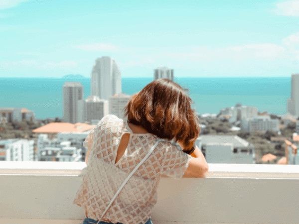 A person leaning over a balcony, overlooking a coastal city with modern buildings and the ocean in the background. The bright, sunny day contrasts with the pensive posture, reflecting the journey of "starting over in your 30s" and the introspection or longing that can come with it.