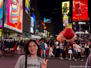 A woman stands in Times Square at night, surrounded by bright lights and bustling city life in Midtown NYC.